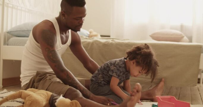 Proud black dad sitting on floor with cute daughter painting on paper