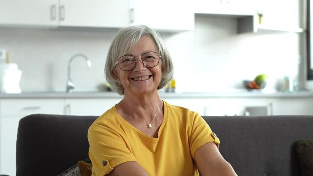 Smiling middle aged mature grey haired woman looking at camera, happy old lady in glasses posing at home indoor, positive single senior retired female sitting on sofa in living room headshot portrait
