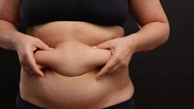 Unrecognizable fat woman wearing sportswear bra panties, standing on black background, squeezing massaging lifting subcutaneous fat of stomach with hands. Self-treatment, body positive, liposuction.