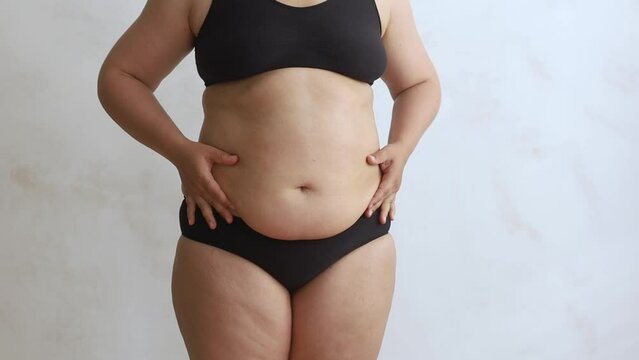 Unrecognizable plump woman wearing black underwear, squeezing excessive subcutaneous fat of stomach on waist, doing massage on white background. Weight loss, body care, plastic surgery, liposuction. 