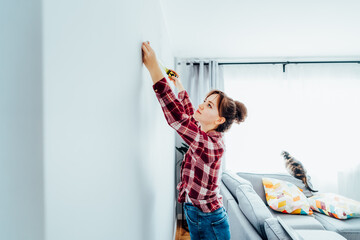 Young woman in plaid shirt doing measuring with a measure tape on the wall. Girl wants to put a...