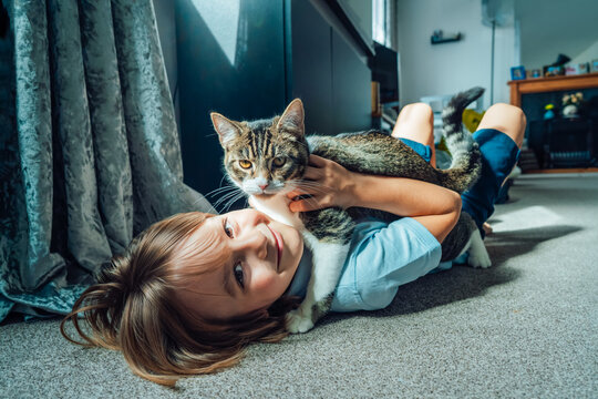 Portrait Of A Happy Smiling Kid At Home Playing With A Kitty Cat. Caucasian Boy Lying On A Carpeted Floor Holding His Cat In The Living Room. Life With A Pet. Best Friends.Selective Focus.