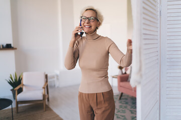 Smiling mature woman answering smartphone and standing in living room