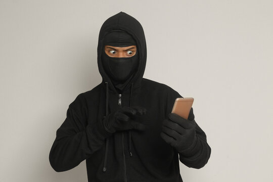 Portrait of mysterious man wearing black hoodie and mask doing hacking activity on mobile phone, hacker holding a smartphone. Isolated image on gray background