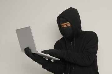 Portrait of mysterious man wearing black hoodie and mask doing hacking activity on laptop, hacker holding a personal computer. Cyber security concept. Isolated image on gray background