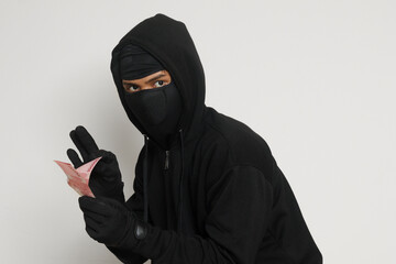 Portrait of mysterious man wearing black hoodie and mask stealing one hundred thousand rupiah successfully from victim. Isolated image on gray background
