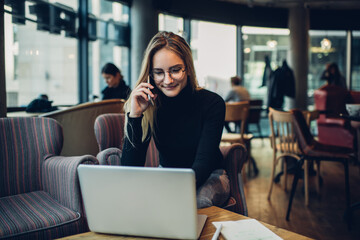 Smiling female freelancer working on laptop in cafe and talking on smartphone