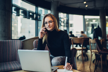 Young female talking on smartphone while working remotely on laptop