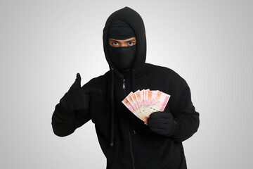 Portrait of mysterious man wearing black hoodie and mask stealing one hundred thousand rupiah successfully from victim. Isolated image on gray background