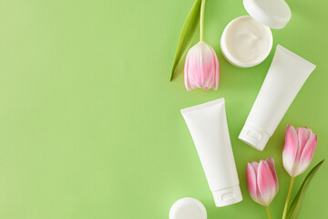 Organic skincare products concept. Top view photo of cosmetic tubes without label open cream jar small hearts baubles and pink tulips on light green background with copy space
