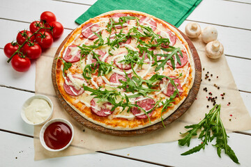 Freshly baked tasty pepperoni pizza with salami, mozzarella cheese and rukkola served on wooden background with tomatoes, sauce and herbs. Food delivery concept. Restaurant menu