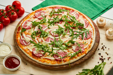 Freshly baked tasty pepperoni pizza with salami, mozzarella cheese and rukkola served on wooden background with tomatoes, sauce and herbs. Food delivery concept. Restaurant menu