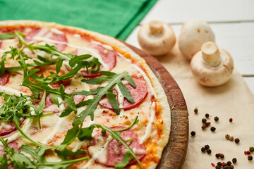 Freshly baked tasty pepperoni pizza with salami, mozzarella cheese and rukkola served on wooden background with tomatoes, sauce and herbs. Food delivery concept. Restaurant menu