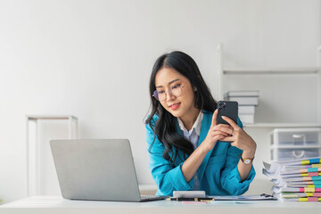 Asian Woman entrepreneur busy with her work in the office. Young Asian woman talking over smartphone or cellphone while working on computer at her desk.