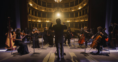 Back View of Professional Conductor Directing Symphony Orchestra with Performers Wearing Medical Masks, Playing Violins, Cello and Trumpet on Classic Theatre with Curtain Stage During Music Concert - obrazy, fototapety, plakaty