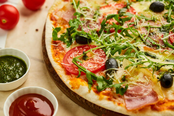 Freshly baked pizza with ham, rukkola, sauce pesto and olives served on wooden background with tomatoes, sauces and herbs. Food delivery concept. Restaurant menu