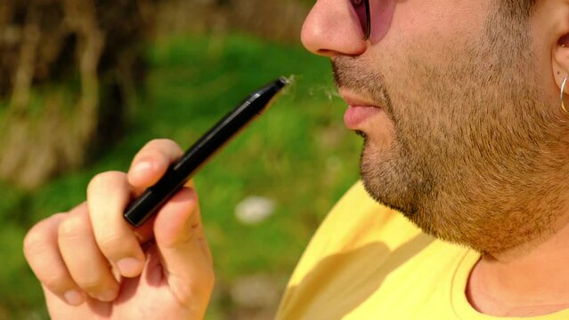 Portrait of a man smoking electronic cigarette in a park. Closeup thoughtful guy with e-cigarette in hand.