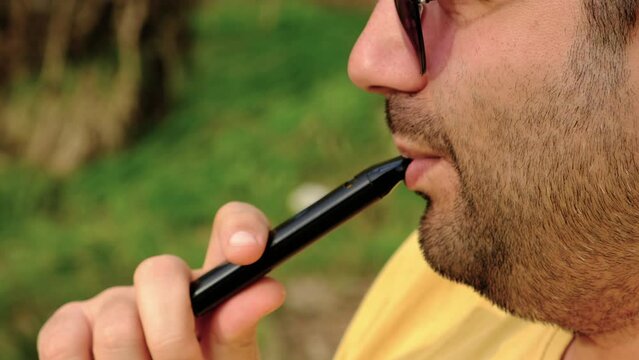 Portrait of a man smoking electronic cigarette in a park. Closeup thoughtful guy with e-cigarette in hand.
