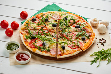 Sliced Freshly baked pizza with ham, rukkola, sauce pesto and olives served on wooden background with tomatoes, sauces and herbs. Food delivery concept. Restaurant menu