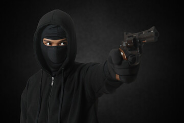 Mysterious man wearing black hoodie and mask holding a pistol, shooting with a gun. Isolated image on dark ambient background