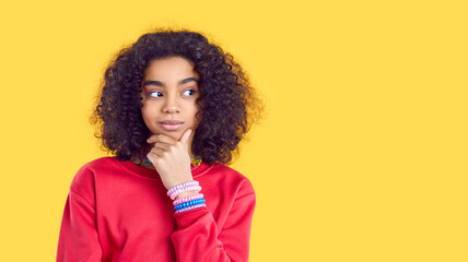Fototapeta na wymiar Curious pretty African American child girl with curly hair in red sweatshirt isolated on yellow color background holding hand on chin, looking to right copy space side and thinking about something