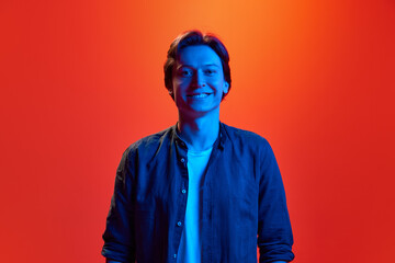 Studio shot of young guy, student smiling broadly over red-orange background in neon light. Wide...