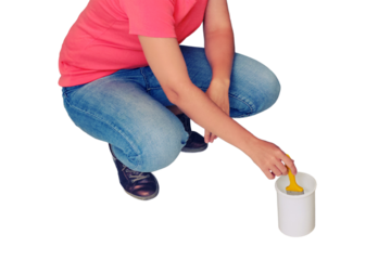 A woman squats and dips a brush into a can of paint, isolated on a white background. Painting the walls yourself in the repair of a new built house.