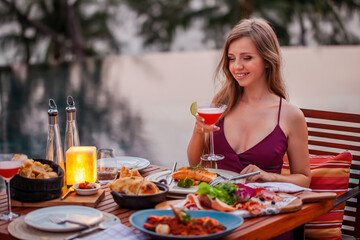Elegant attractive smiling woman on romantic dinner in luxury gourmet restaurant. Happy young lady with a glass of cocktail in fine dining outdoor restaurant. Sexy elegant girl celebrating holiday.