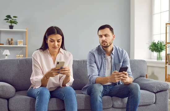 Jealous man spying looking at his girlfriend phone. Couple sitting on couch with smartphones, suspicious husband trying to read messages at wife smartphone while she texting message and smiling