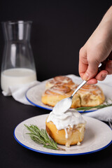 cinnamon rolls on white plate with blue rim decorated with rosemary and jug of milk. The hand holding a spoon with cream