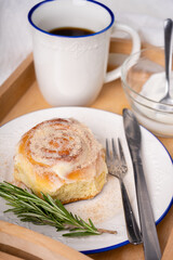 cinnamon roll with rosemary on a white plate, a cup of coffee and cream on a wooden tray