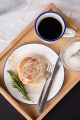 cinnamon roll with rosemary on a white plate, a cup of coffee and cream on a wooden tray
