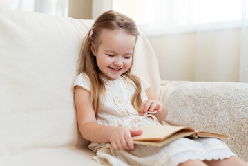 Obraz na płótnie Canvas Little cute blond girl reading book siting on a sofa. Child reading, dreaming and imagination development. International Children's Book Day. Kids Love to reading. World Book Day