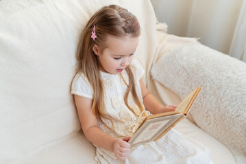 Little cute blond girl reading book siting on a sofa. Child reading, dreaming and imagination development. International Children's Book Day. Kids Love to reading. World Book Day