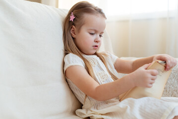 Little cute blond girl reading book siting on a sofa. International Children's Book Day. Kids Love to reading. World Book Day. Child reading, dreaming and imagination development.