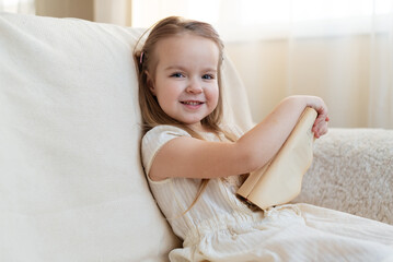 Obraz na płótnie Canvas Little cute blond girl reading book siting on a sofa. International Children's Book Day. Kids Love to reading. World Book Day. Child reading, dreaming and imagination development. 