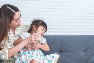 Asian mother holding a glass of milk to feed her 3-year-old daughter who refuses to drink milk....