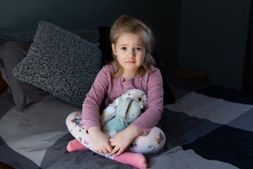 Adorable fair little girl with sad expression sitting cross-legged on bed holding plush toy rabbits
