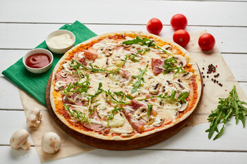Freshly baked pizza with ham, rukkola and mushrooms served on wooden background with tomatoes, sauces and herbs. Food delivery concept. Restaurant menu