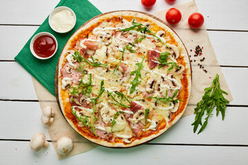 Freshly baked pizza with ham, rukkola and mushrooms served on wooden background with tomatoes, sauces and herbs. Food delivery concept. Restaurant menu. Top view
