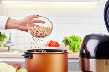 Female hand pours peas into a multi cooker. Multi-cooker among of various raw foods on kitchen table. Woman preparing seed or cereal in a slow cooker.