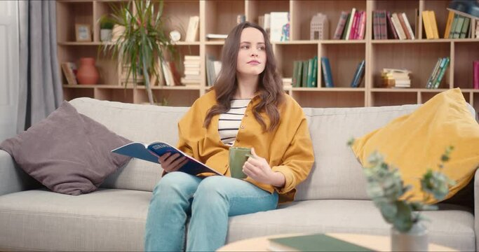 Beautiful Caucasian woman relaxing in library. Pretty focused female reading magazine and drinking tea or coffee. After closing journal, thinking about her plans for next purchase.