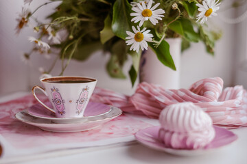 Fototapeta na wymiar A gorgeous fragile white-pink coffee cup on a large soft pink napkin and a puffy airy pink marshmallow in a saucer on a white table. Stylish polka dot scarf and beautiful sunny daisies in white vase.