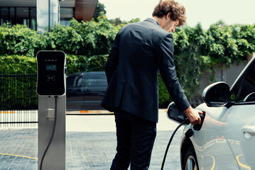 Progressive businessman with electric car recharging at public charging station at modern city...