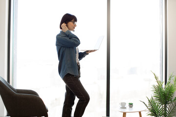 Charming female employee typing on wireless laptop while standing near panoramic window at office. Dark haired woman in casual outfit and glasses checking emails on modern gadget at workplace.