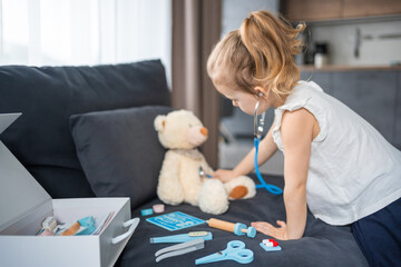 Little girl playing doctor with toys and teddy bear on the sofa in living room at home