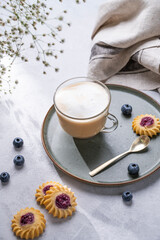Cappuccino or latte with milk foam in a cup with homemade berry cookies and blueberries on a light background with gypsophila branches. Concept spring morning breakfast.