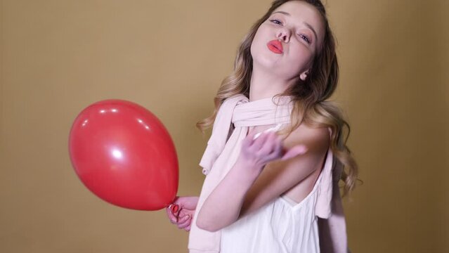 Portrait of excited young girl dancing in trendy summer white dress. Woman model with red air balloon posing on golden background. Ready for party. Slow motion