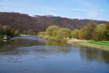 Flusslandschaft mit Spiegelung von Bäumen in Wasser, grüner Wiese mit Bäumen am Ufer vor Berghügel mit Wald und blauem Himmel bei Sonne am Mittag im Frühling 