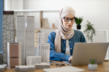 Focused bespectacled muslim female in hijab typing on portable computer at office desk with building models on it. Experienced architect developing urban design using modern gadgets and drawing tools.